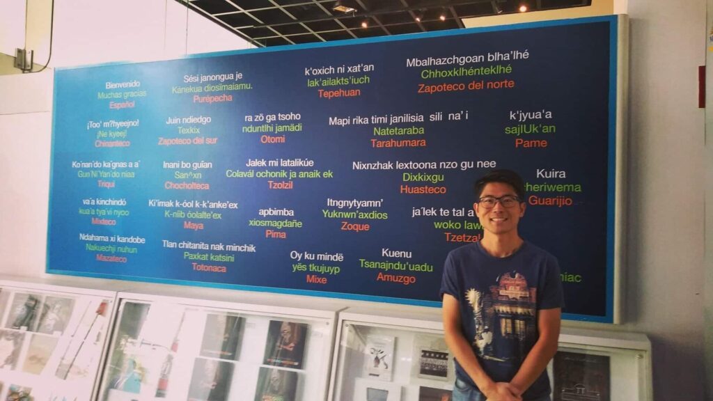 メキシコの観光ガイド岩﨑功がいろいろな言語が書かれたボードの前で挨拶をしている